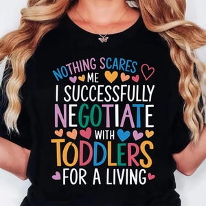 May include: Black t-shirt with the text "NOTHING SCARES ME I SUCCESSFULLY NEGOTIATE WITH TODDLERS FOR A LIVING" in colorful letters and heart graphics. The shirt is a crew neck.