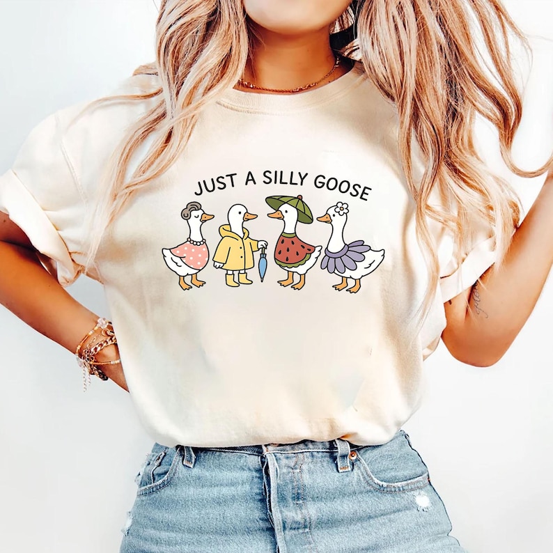 May include: Cream-colored t-shirt featuring the phrase "JUST A SILLY GOOSE" above a cartoon illustration of four geese. Each goose is uniquely dressed, including a pink dress, yellow raincoat, watermelon-themed outfit, and a lavender skirt.
