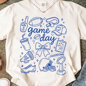 Puede incluir: Camiseta de color crema con un diseño gráfico azul que presenta iconos relacionados con el fútbol y las palabras "game day" y "touchdown". El diseño incluye un balón de fútbol, un casco y otros elementos deportivos.