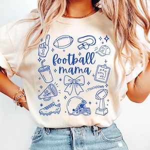 Puede incluir: Sudadera color crema con un diseño gráfico azul. El diseño incluye iconos relacionados con el fútbol como un balón, un casco y un trofeo, junto con las palabras "football mama". El diseño está rodeado de estrellas y otros elementos decorativos.