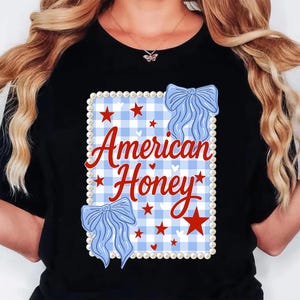 May include: Black t-shirt with a design that reads "American Honey" in red script. The design includes a blue and white gingham pattern, red stars, and blue bow accents, framed by a pearl border.