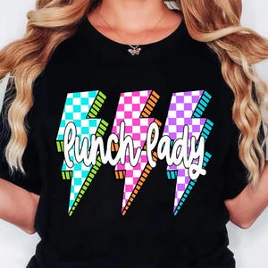 K&ouml;nnte beinhalten: Schwarzes T-Shirt mit drei Blitz-Grafiken in t&uuml;rkis, rosa und lila Karomustern. Die Worte "Lunch Lady" sind in wei&szlig;er Schrift &uuml;ber den Blitzen geschrieben.