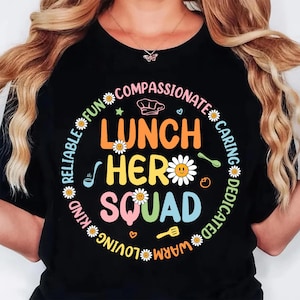 K&ouml;nnte beinhalten: Schwarzes T-Shirt mit einem farbenfrohen Aufdruck &bdquo;Lunch Hero Squad&ldquo;. Das Design umfasst G&auml;nsebl&uuml;mchen, eine Kochm&uuml;tze und W&ouml;rter wie &bdquo;mitf&uuml;hlend&ldquo; und &bdquo;f&uuml;rsorglich&ldquo; in einem kreisf&ouml;rmigen Muster um den zentralen Text.