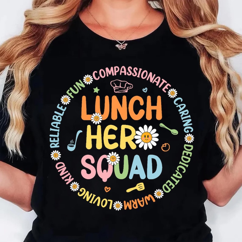 K&ouml;nnte beinhalten: Schwarzes T-Shirt mit einem farbenfrohen Aufdruck &bdquo;Lunch Hero Squad&ldquo;. Das Design umfasst G&auml;nsebl&uuml;mchen, eine Kochm&uuml;tze und W&ouml;rter wie &bdquo;mitf&uuml;hlend&ldquo; und &bdquo;f&uuml;rsorglich&ldquo; in einem kreisf&ouml;rmigen Muster um den zentralen Text.