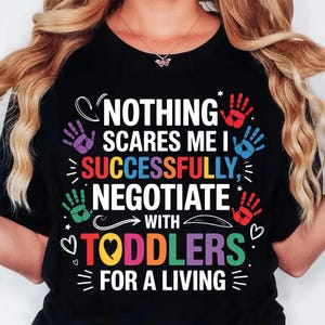 May include: Black t-shirt with the text "NOTHING SCARES ME I SUCCESSFULLY NEGOTIATE WITH TODDLERS FOR A LIVING." The design includes colorful handprints and hearts.
