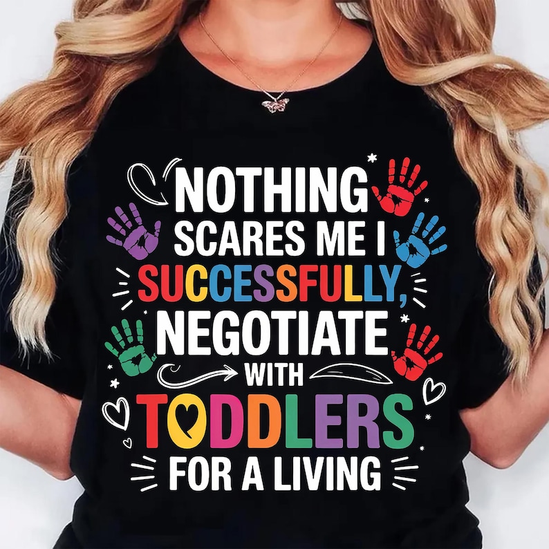 May include: Black t-shirt with the text "NOTHING SCARES ME I SUCCESSFULLY NEGOTIATE WITH TODDLERS FOR A LIVING." The design includes colorful handprints and hearts.