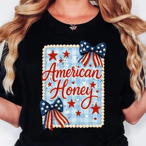 May include: Black t-shirt with a patriotic design. The design features the text "American Honey" in red script, set against a blue and white plaid background. Red stars and blue bows with white stars and red stripes complete the design.