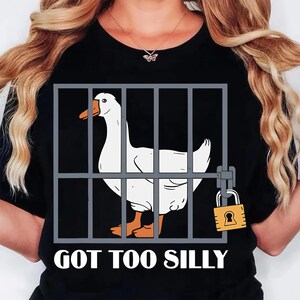 Może przedstawiać: Czarny t-shirt z białym gęsią w szarej klatce, zabezpieczonej ż&oacute;łtą kł&oacute;dką. Gęś ma pomarańczowe stopy i dzi&oacute;b. Tekst "GOT TOO SILLY" jest wydrukowany na dole w kolorze białym.