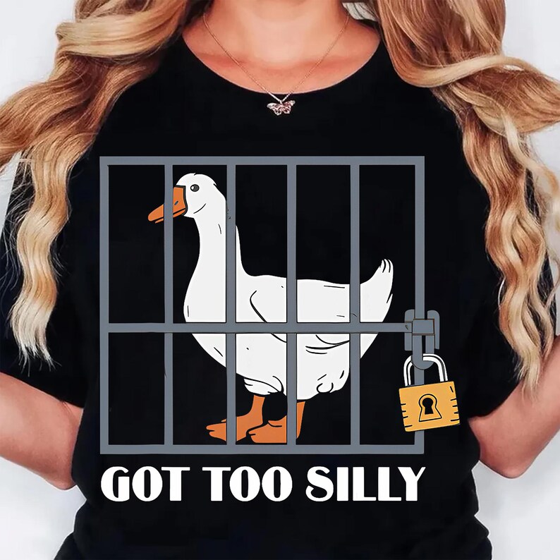 Może przedstawiać: Czarny t-shirt z białym gęsią w szarej klatce, zabezpieczonej ż&oacute;łtą kł&oacute;dką. Gęś ma pomarańczowe stopy i dzi&oacute;b. Tekst "GOT TOO SILLY" jest wydrukowany na dole w kolorze białym.