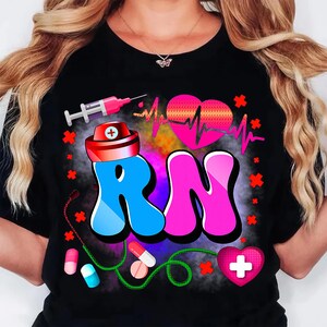 Pu&ograve; includere: T-shirt nero con un design grafico colorato. Il design include le lettere "RN" in blu e rosa, un cappello da infermiera rosso e bianco, un cuore rosa con una linea di battito cardiaco, una siringa e pillole mediche.