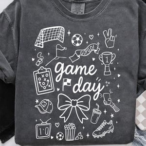 Puede incluir: Sudadera gris oscuro con elementos gr&aacute;ficos blancos relacionados con el f&uacute;tbol. El dise&ntilde;o incluye una porter&iacute;a de f&uacute;tbol, un bal&oacute;n, un trofeo y las palabras "game day". Otras ilustraciones incluyen un silbato, un lazo y palomitas de ma&iacute;z.