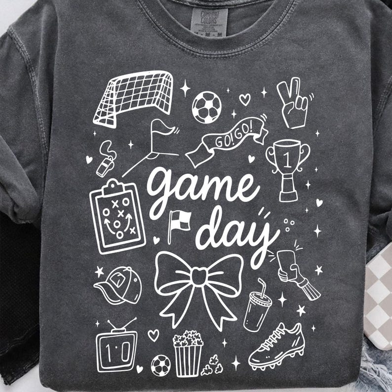 Puede incluir: Sudadera gris oscuro con elementos gr&aacute;ficos blancos relacionados con el f&uacute;tbol. El dise&ntilde;o incluye una porter&iacute;a de f&uacute;tbol, un bal&oacute;n, un trofeo y las palabras "game day". Otras ilustraciones incluyen un silbato, un lazo y palomitas de ma&iacute;z.
