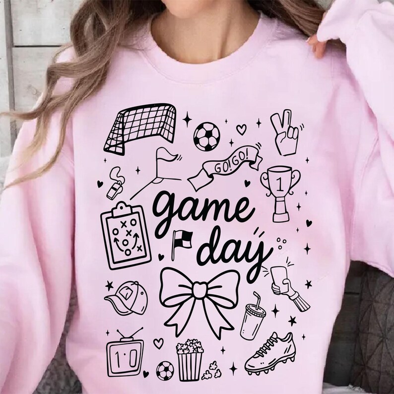 Puede incluir: Sudadera rosa claro con un dise&ntilde;o gr&aacute;fico negro con ilustraciones tem&aacute;ticas de f&uacute;tbol y las palabras "game day". El dise&ntilde;o incluye una red de f&uacute;tbol, un bal&oacute;n, un trofeo y otros iconos deportivos.