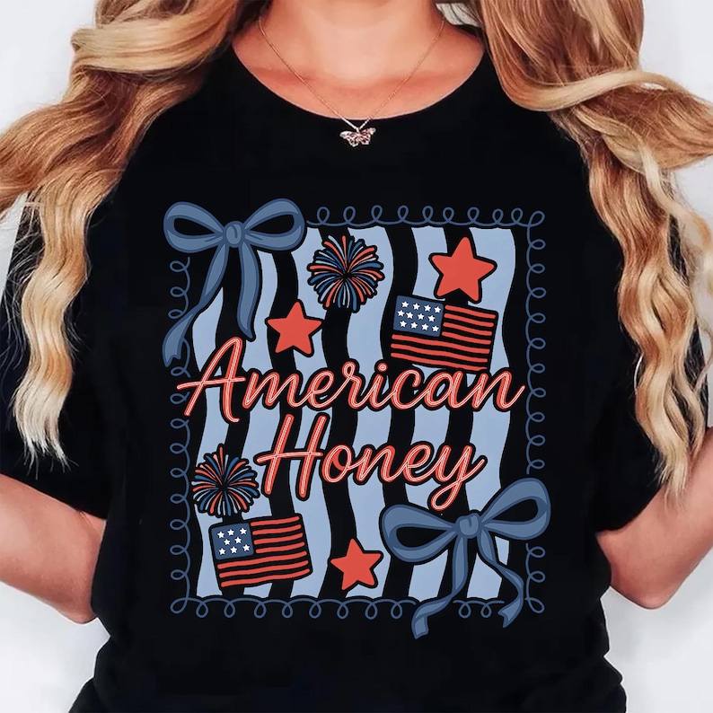 May include: Black t-shirt with a patriotic design. The design features the text "American Honey" in red and blue, along with red stars, blue bows, fireworks, and American flags. The shirt is a crew neck.