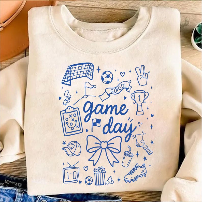 Puede incluir: Sudadera de color crema con un dise&ntilde;o gr&aacute;fico azul que presenta garabatos con tem&aacute;tica de f&uacute;tbol y las palabras "game day". El dise&ntilde;o incluye una red de f&uacute;tbol, un bal&oacute;n, un trofeo y otros iconos relacionados con el deporte. La sudadera es de material suave.