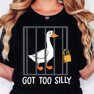 Może przedstawiać: Czarny t-shirt z kresk&oacute;wkową gęsią w klatce, zabezpieczoną kł&oacute;dką. Gęś jest biała z pomarańczowymi stopami i dziobem. Tekst "GOT TOO SILLY" jest wydrukowany poniżej w kolorze białym.