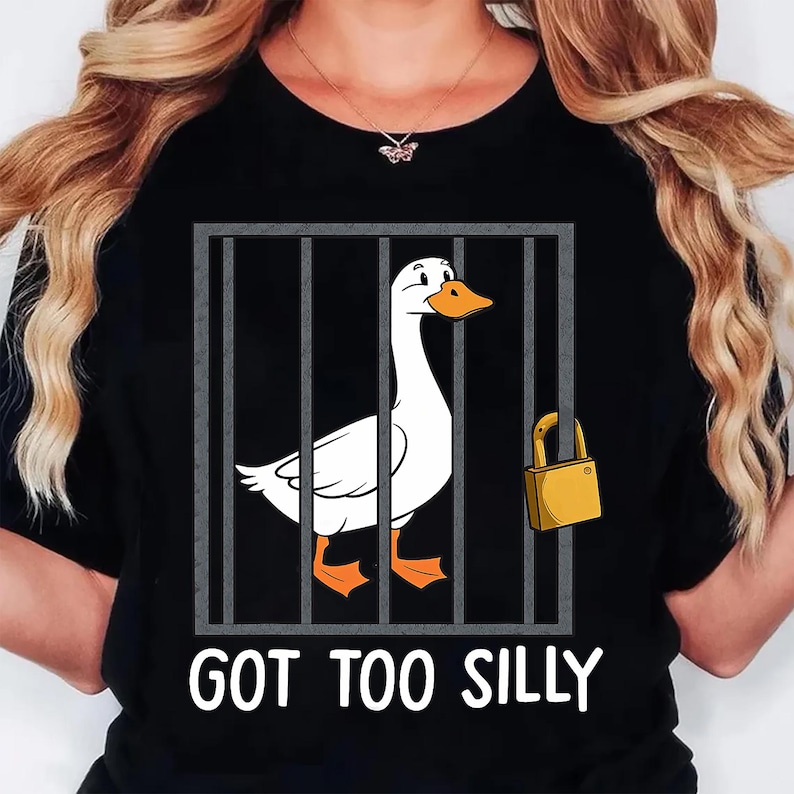 Może przedstawiać: Czarny t-shirt z kresk&oacute;wkową gęsią w klatce, zabezpieczoną kł&oacute;dką. Gęś jest biała z pomarańczowymi stopami i dziobem. Tekst "GOT TOO SILLY" jest wydrukowany poniżej w kolorze białym.