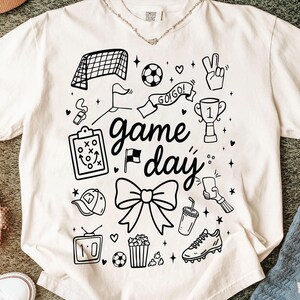 Puede incluir: Camiseta de color crema con un dise&ntilde;o gr&aacute;fico negro con elementos relacionados con el f&uacute;tbol. El dise&ntilde;o incluye una red de porter&iacute;a, un bal&oacute;n, un trofeo y las palabras "game day". Otros elementos incluyen un lazo, palomitas de ma&iacute;z y una bota de f&uacute;tbol.