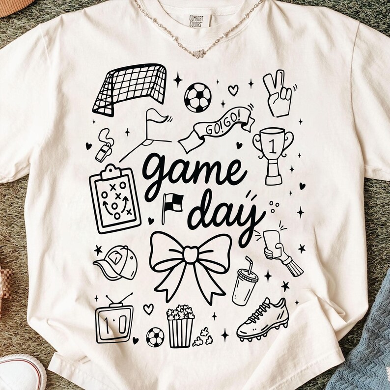 Puede incluir: Camiseta de color crema con un dise&ntilde;o gr&aacute;fico negro con elementos relacionados con el f&uacute;tbol. El dise&ntilde;o incluye una red de porter&iacute;a, un bal&oacute;n, un trofeo y las palabras "game day". Otros elementos incluyen un lazo, palomitas de ma&iacute;z y una bota de f&uacute;tbol.