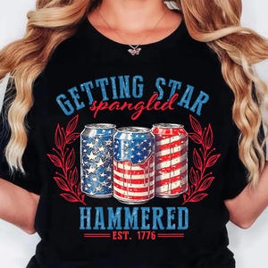 May include: Black t-shirt with a patriotic design featuring three beverage cans decorated with the American flag. The text "GETTING STAR spangled HAMMERED EST. 1776" is printed above and below the cans in blue and red.