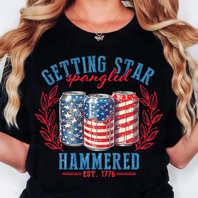 May include: Black t-shirt with a patriotic design featuring three beverage cans decorated with the American flag. The text "GETTING STAR spangled HAMMERED EST. 1776" is printed above and below the cans in blue and red.