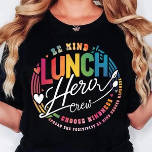 K&ouml;nnte beinhalten: Schwarzes T-Shirt mit einem farbenfrohen Grafikdesign. Das Design enth&auml;lt die Worte "BE KIND LUNCH Hero crew CHOOSE KINDNESS" in einem regenbogenfarbenen Kreis mit Herzen und einem Regenbogen. Der Satz "SPREAD THE POSITIVITY BE KIND" ist ebenfalls enthalten.