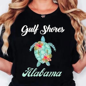 May include: Black t-shirt featuring the text "Gulf Shores Alabama" in white and a turquoise sea turtle design with floral accents. The turtle has a light blue shell and is decorated with colorful flowers.