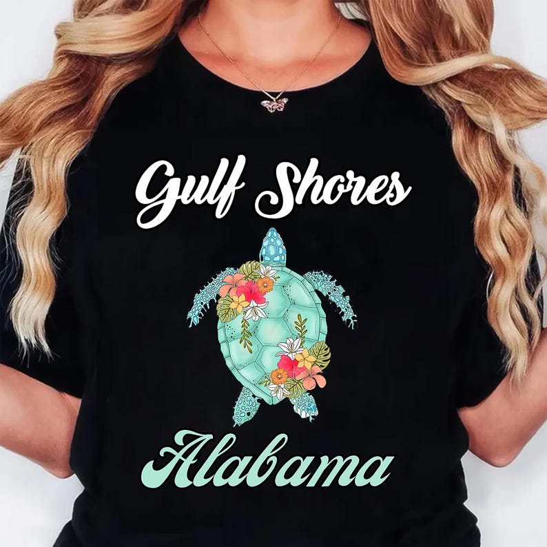 May include: Black t-shirt featuring the text "Gulf Shores Alabama" in white and a turquoise sea turtle design with floral accents. The turtle has a light blue shell and is decorated with colorful flowers.
