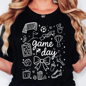 Puede incluir: Camiseta negra con un dise&ntilde;o gr&aacute;fico blanco que presenta ilustraciones con tem&aacute;tica de f&uacute;tbol y las palabras "game day". El dise&ntilde;o incluye una porter&iacute;a de f&uacute;tbol, un bal&oacute;n, un trofeo y otros iconos relacionados. La camiseta es de estilo casual.