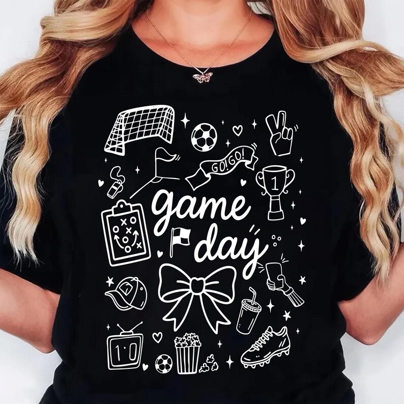 Puede incluir: Camiseta negra con un dise&ntilde;o gr&aacute;fico blanco que presenta ilustraciones con tem&aacute;tica de f&uacute;tbol y las palabras "game day". El dise&ntilde;o incluye una porter&iacute;a de f&uacute;tbol, un bal&oacute;n, un trofeo y otros iconos relacionados. La camiseta es de estilo casual.