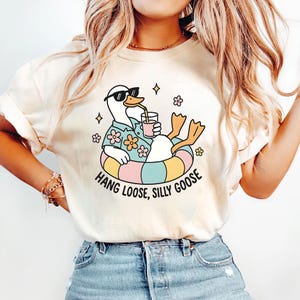 May include: Cream-colored t-shirt featuring a cartoon goose wearing sunglasses, a Hawaiian shirt, and an inner tube, holding a drink. The text "Hang Loose, Silly Goose" is printed below the goose in a playful font. The shirt has a relaxed fit.