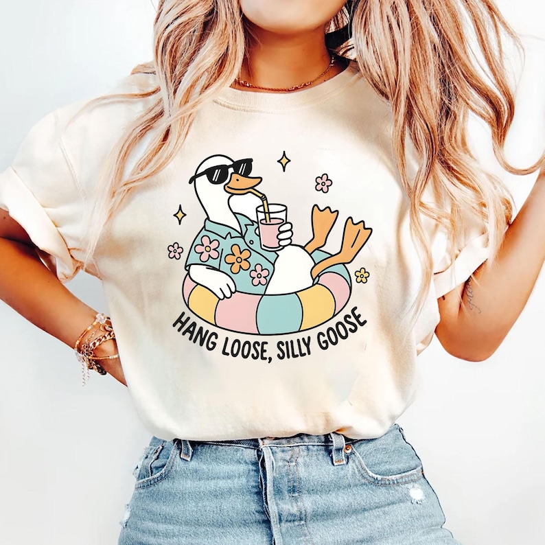 May include: Cream-colored t-shirt featuring a cartoon goose wearing sunglasses, a Hawaiian shirt, and an inner tube, holding a drink. The text "Hang Loose, Silly Goose" is printed below the goose in a playful font. The shirt has a relaxed fit.