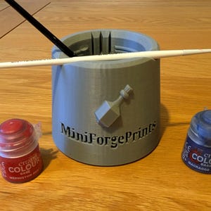 Può includere: Un portapennelli grigio stampato in 3D con la scritta "MiniForgePrints" e un disegno di bottiglia. Sono visibili anche due pennelli e due vasetti di vernice, uno rosso e uno blu.