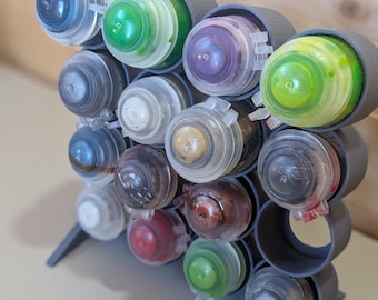 Puede incluir: Un soporte de plástico gris para pintura que contiene quince botes de pintura circulares pequeños con tapas transparentes abatibles. Los botes contienen varios colores, incluyendo verde, azul, morado, blanco, oro, marrón y negro.