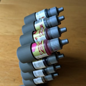 May include: A gray 3D-printed paint bottle holder with several small paint bottles. The bottles are filled with various colors of paint, including red, white, and brown. The holder is designed to keep the bottles organized and accessible.