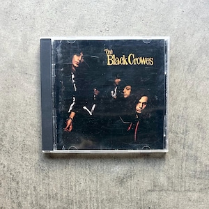 May include: A CD case featuring the album cover for "The Black Crowes." The cover art shows four men in dark clothing against a black background. The band's name is in gold lettering.