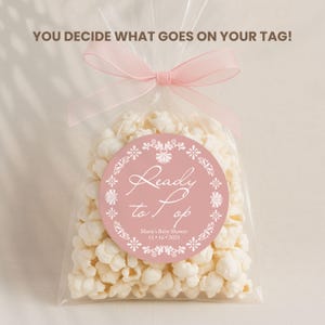 May include: A clear cellophane bag filled with popcorn, tied with a pink ribbon. A round, pink tag with white floral accents reads "Ready to Pop" with event details. The text "YOU DECIDE WHAT GOES ON YOUR TAG!" is at the top.