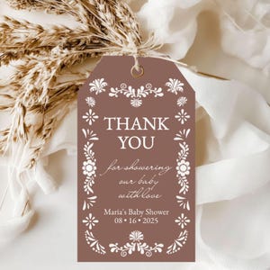 May include: A brown tag with white floral designs and the words "THANK YOU" in bold letters. The tag also includes the text "for showering our baby with love" and "Maria's Baby Shower 08.16.2025".
