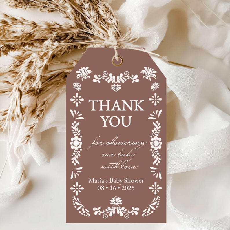May include: A brown tag with white floral designs and the words "THANK YOU" in bold letters. The tag also includes the text "for showering our baby with love" and "Maria's Baby Shower 08.16.2025".