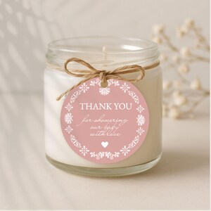May include: A clear glass jar candle with a white wax interior. A pink tag with white floral accents and the text "THANK YOU for showering our baby with love" is tied around the jar with twine.