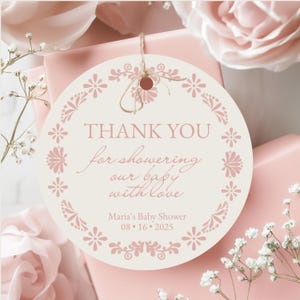 May include: A round, cream-colored baby shower gift tag with the words "THANK YOU for showering our baby with love" in pink script. The tag is decorated with pink floral designs and the text "Maria's Baby Shower 08.16.2025". The tag is tied with twine.