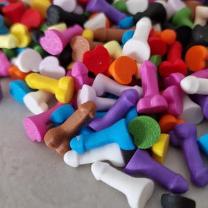 Op de afbeelding: Een kleurrijk assortiment kleine, fallusvormige objecten in verschillende kleuren, waaronder roze, blauw, geel, bruin en wit. Sommige zijn hartvormig. De objecten zijn verspreid, wat een speelse en levendige weergave creëert.