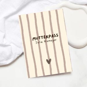 Könnte beinhalten: Ein rechteckiges Heft mit cremefarbenem Einband und vertikalen taupefarbenen Streifen. Der Text MUTTERPASS und Julia Niemeyer ist in Dunkelbraun gedruckt, ergänzt durch ein kleines dunkelbraunes Herzsymbol nahe der unteren Kante.
