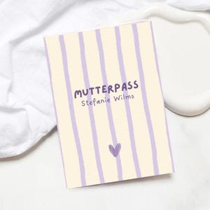 Könnte beinhalten: Ein rechteckiges Notizbuch mit cremefarbenem Hintergrund und vertikalen lavendelfarbenen Streifen. Auf dem Cover steht das Wort "MUTTERPASS" in Lila, zusammen mit dem Namen "Stefanie Wilms" und einem kleinen lila Herz.