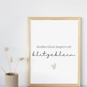 May include: A framed print with the German text "Großes Glück beginnt oft klitzeklein" and a small heart illustration. The frame is light wood, and a beige vase with dried flowers sits to the left.