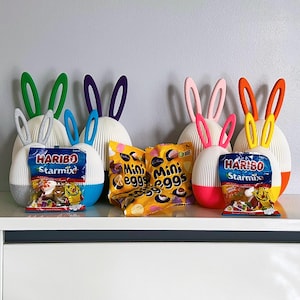 Op de afbeelding: Vier witte paaseieren in de vorm van een konijn met kleurrijke oren. De containers zijn gevuld met snoep. Twee zakjes mini-eieren zijn voor de containers. De zakjes zijn gelabeld met "Mini Eggs". Twee zakjes Haribo Starmix snoep zijn voor de containers. De zakjes zijn gelabeld met "Haribo Starmix".