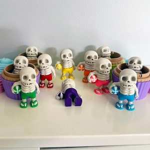 Può includere: Una collezione di figurine di scheletri stravaganti in abbigliamento da spiaggia, che tengono palle da spiaggia, e alcuni sono in contenitori a forma di cupcake. Gli scheletri indossano pantaloncini e sandali colorati. Forme decorative a spirale rosa e blu sui lati.