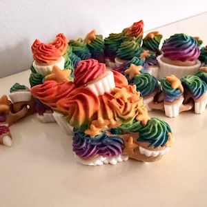 Puede incluir: Una figura de dragón articulada y flexible construida con segmentos interconectados que parecen cupcakes con bases blancas y glaseado en espiral de colores arcoíris. La criatura presenta pequeños detalles en forma de estrella y garras blancas en sus extremidades.