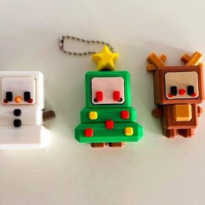 Könnte beinhalten: Drei Schlüsselanhänger zum Thema Weihnachten: ein weißer Schneemann, ein grüner Weihnachtsbaum mit gelbem Stern und ein braunes Rentier. Jeder Schlüsselanhänger hat ein pixeliges Design mit schwarzen, roten und braunen Akzenten. Der Baum hat eine silberne Kette.