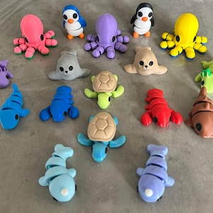 Op de afbeelding: Een verzameling kleurrijke miniatuurzeedierenbeeldjes. De collectie omvat pinguïns, octopussen, zeehonden, schildpadden, kreeften en vissen in verschillende tinten blauw, roze, geel, groen en bruin. Deze kleine, gedetailleerde figuren zijn gerangschikt op een neutrale ondergrond.